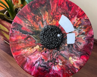 May include: A circular red, black, white, and gold abstract painting with a black textured center. The painting is on a wooden easel.