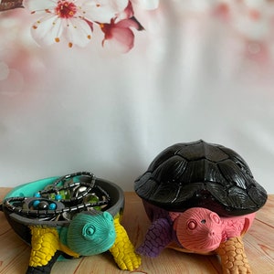 Puede incluir: Dos platos para joyas con forma de tortuga, uno con una concha negra y detalles en rosa, morado y naranja, y el otro con una concha turquesa y detalles en amarillo, negro y gris. Ambos platos están llenos de joyas.