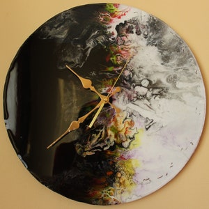 May include: Round wall clock with a unique abstract design. The clock face features a glossy black and white marbled pattern with gold-colored hands and a central mechanism. The design incorporates splashes of color.