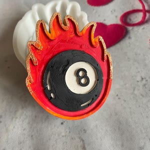 Op de afbeelding: Een rode en oranje siliconen mal in de vorm van een brandende achtbal. De mal is gedetailleerd met een gouden omtrek en een zwarte achtbal met een wit centrum.