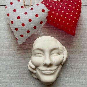 May include: White ceramic sculpture of a smiling face with a hand on the cheek. The sculpture is surrounded by two red and white polka dot hearts.
