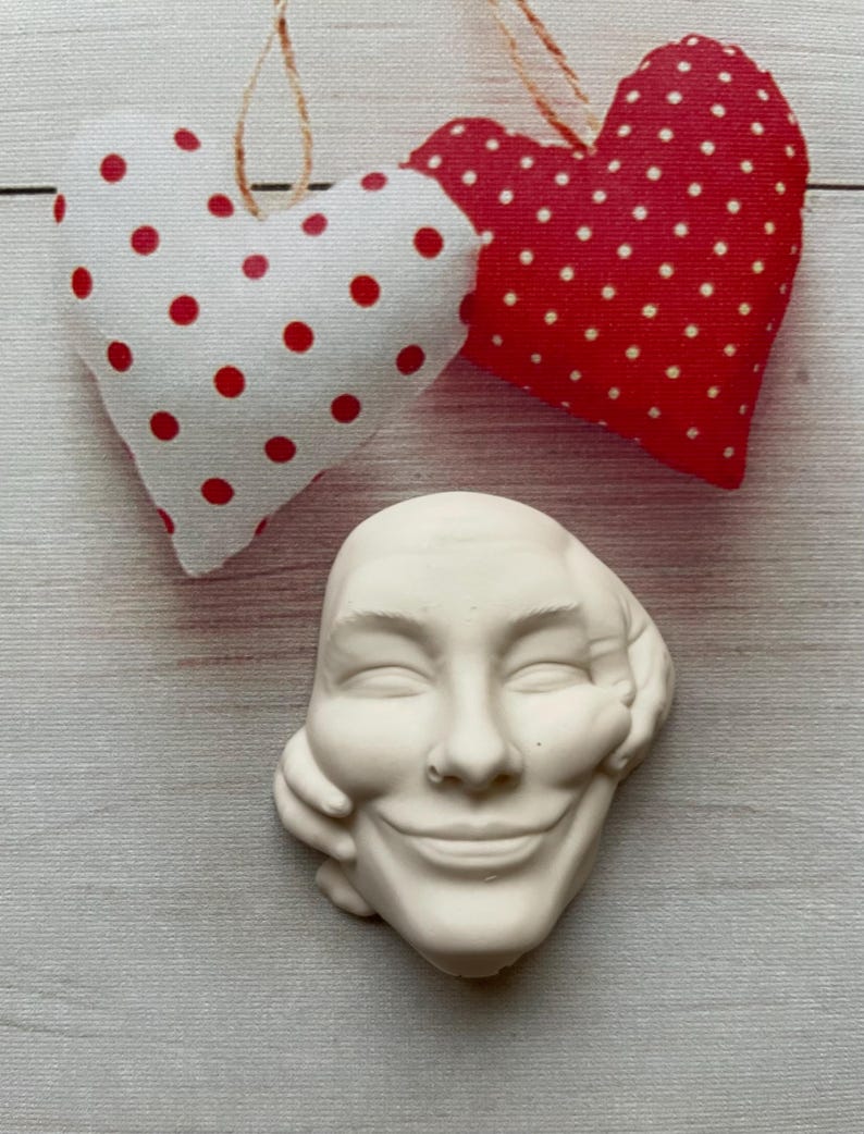 May include: White ceramic sculpture of a smiling face with a hand on the cheek. The sculpture is surrounded by two red and white polka dot hearts.