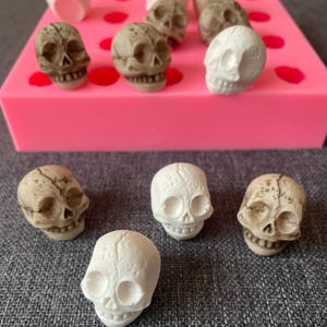 May include: A collection of small skull figurines in various shades of white and tan. Some skulls are arranged on a pink mold with red circles. The skulls have detailed facial features, including eye sockets and teeth. The background is a gray surface.