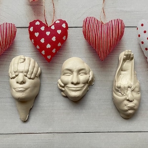 May include: Three white plaster sculptures of faces, each with a hand covering one of the senses. The sculptures are arranged in a row on a white wooden surface. Above the sculptures are three red and white fabric hearts hanging from strings.