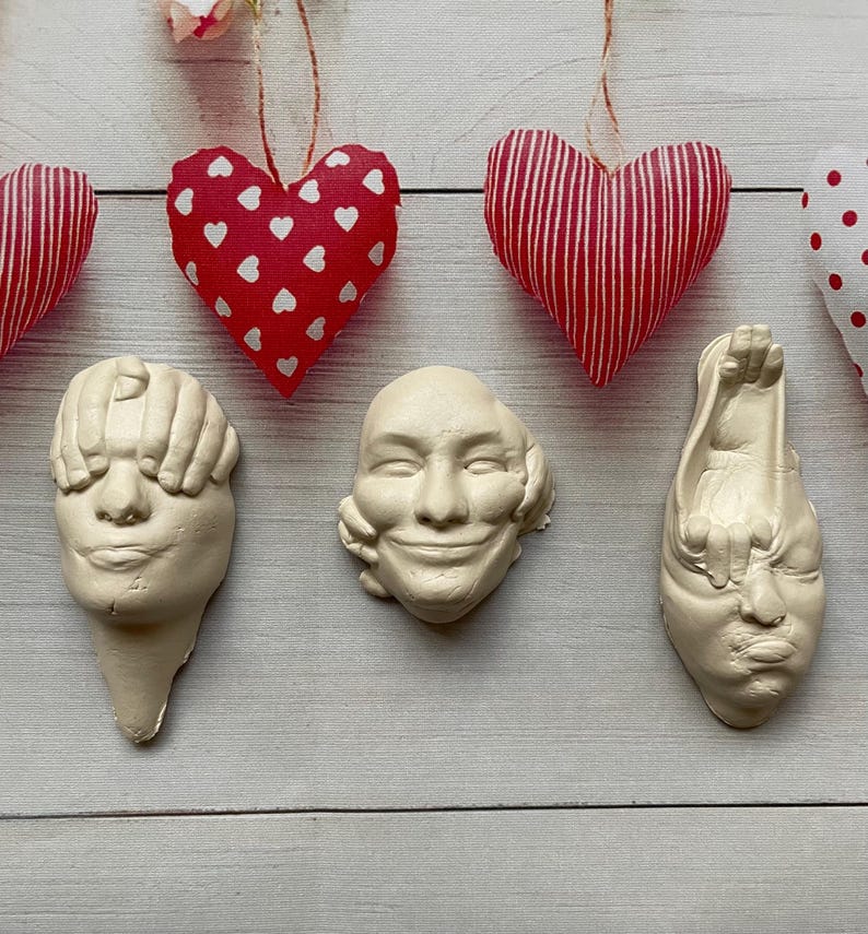May include: Three white plaster sculptures of faces, each with a hand covering one of the senses. The sculptures are arranged in a row on a white wooden surface. Above the sculptures are three red and white fabric hearts hanging from strings.