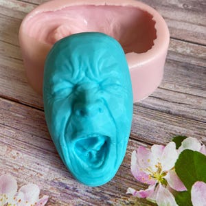 Puede incluir: Un molde de silicona azul de una cara gritando. El molde está sentado sobre una superficie de madera con flores rosas.