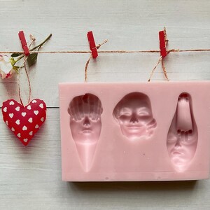 May include: Pink silicone mold with three face-shaped cavities. The mold is for making chocolate or other edible treats.