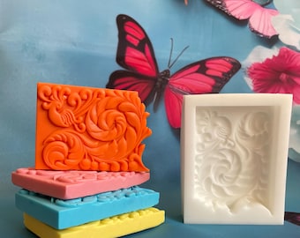 Könnte beinhalten: Eine weiße Silikonform zum Herstellen von Seife oder anderen Bastelarbeiten. Die Form ist quadratisch mit einem detaillierten floralen Design. Es gibt vier orangefarbene Seifenstücke, die übereinander gestapelt sind, jedes mit dem gleichen floralen Design.