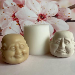 May include: Three small, white ceramic figurines of Buddha faces with different expressions. The faces are smiling, frowning, and neutral. The figurines are sitting on a white surface.