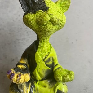 May include: A green and black cat figurine sitting in a meditative pose with its eyes closed. The cat is wearing a black and green robe and has a yellow and purple paw.