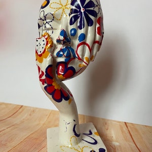 May include: White ceramic sculpture of a face with hands covering the eyes. The sculpture is decorated with colourful flowers in shades of red, blue, yellow, and white.
