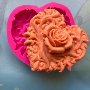 May include: Pink silicone mold in the shape of a heart with a detailed rose and floral design. The mold is filled with a light orange colored resin.