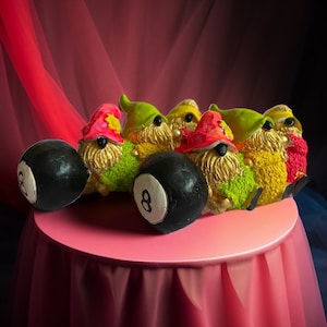 May include: Five gnomes wearing colorful hats and holding black 8-ball pool balls. The gnomes are sitting on a pink table with a red tablecloth.