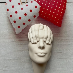 May include: White ceramic sculpture of a face with hands covering the eyes. The sculpture is mounted on a white wall with two red and white polka dot hearts hanging above it.