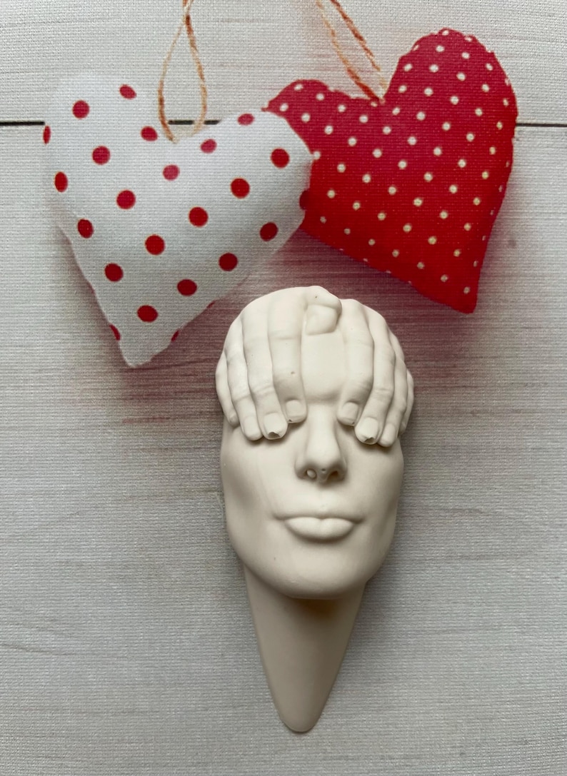 May include: White ceramic sculpture of a face with hands covering the eyes. The sculpture is mounted on a white wall with two red and white polka dot hearts hanging above it.