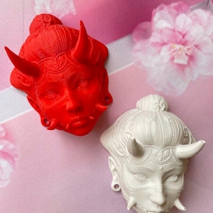 Puede incluir: Dos máscaras decorativas, una roja y otra blanca, cada una representando una cara estilizada con cuernos y detalles intrincados. Las máscaras se colocan sobre un fondo floral rosa, creando un contraste llamativo.