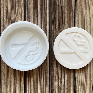 Puede incluir: Molde de silicona blanco para hacer una señal de prohibido fumar. El molde tiene forma de círculo con una línea diagonal que lo atraviesa y un cigarrillo con humo saliendo de él.