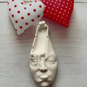 May include: White ceramic sculpture of a face with a hand reaching out from the side of the head. The sculpture has a minimalist style and is a unique home decor piece.