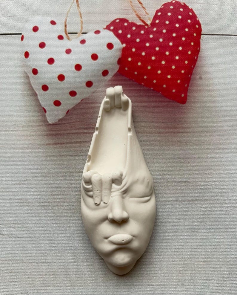 May include: White ceramic sculpture of a face with a hand reaching out from the side of the head. The sculpture has a minimalist style and is a unique home decor piece.