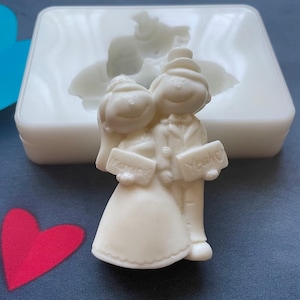 May include: White silicone mold for making a wedding cake topper. The mold features a bride and groom holding signs that say "Marry Me" and "I Do".