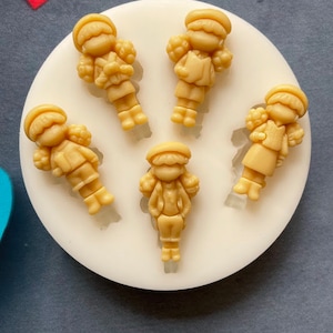 Puede incluir: Cinco pequeñas figuritas detalladas de color beige que representan a niños con sombreros y sosteniendo objetos. Las figuritas están dispuestas en un molde de silicona circular blanco.