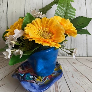 Puede incluir: Un ramo de flores amarillas y blancas en una maceta de cerámica azul con forma de cara. La maceta está sobre un platillo de cerámica azul y blanco.