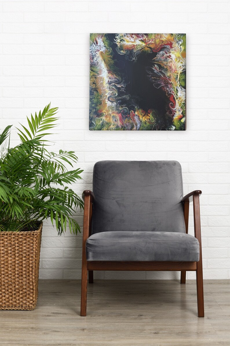 Pode incluir: Pintura abstrata com cores que giram em verde, amarelo, vermelho e branco em torno de um centro preto. A pintura est&aacute; pendurada em uma parede de tijolo branco acima de uma poltrona cinza com pernas de madeira.