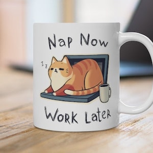 May include: White ceramic mug with a cartoon illustration of a sleeping orange tabby cat on a laptop screen, accompanied by a small coffee cup. The text "Nap Now Work Later" is printed on the mug.