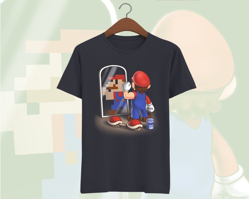 May include: A navy blue t-shirt with a graphic of a pixelated Super Mario character looking in a mirror at a more realistic version of himself. The realistic Mario is wearing a red cap, blue overalls, and white gloves. He is holding a paintbrush and a can of paint.