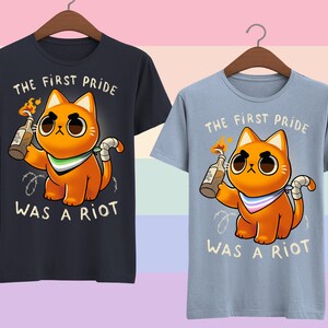 Puede incluir: Dos camisetas con un gr&aacute;fico de un gato naranja sosteniendo un c&oacute;ctel Molotov. El texto en las camisetas dice "The first pride was a riot".