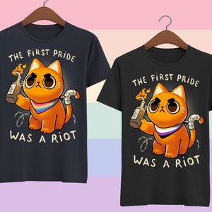Puede incluir: Dos camisetas negras con un gr&aacute;fico de un gato naranja sosteniendo un c&oacute;ctel Molotov. El gato lleva una bandana con los colores de la bandera del orgullo asexual. El texto en las camisetas dice "The First Pride Was A Riot."