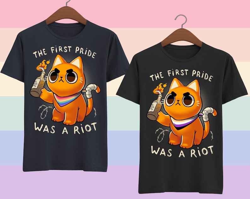 Puede incluir: Dos camisetas negras con un gr&aacute;fico de un gato naranja sosteniendo un c&oacute;ctel Molotov. El gato lleva una bandana con los colores de la bandera del orgullo asexual. El texto en las camisetas dice "The First Pride Was A Riot."