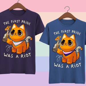 Puede incluir: Dos camisetas azules con un gato naranja de dibujos animados sosteniendo un c&oacute;ctel Molotov. El gato lleva una bandana de orgullo transg&eacute;nero. El texto en las camisetas dice "The First Pride Was A Riot".