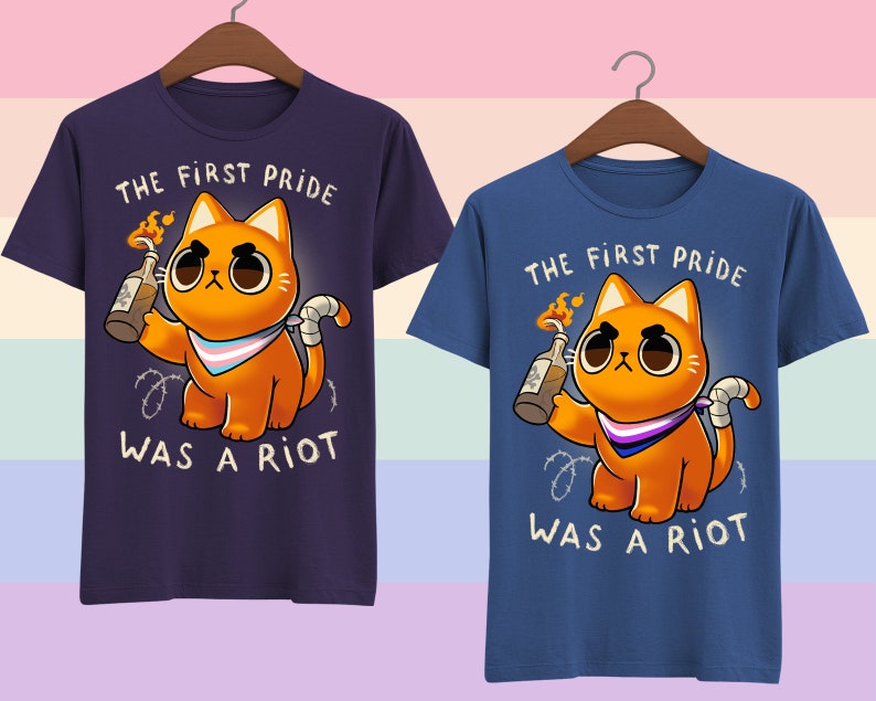 Puede incluir: Dos camisetas azules con un gato naranja de dibujos animados sosteniendo un c&oacute;ctel Molotov. El gato lleva una bandana de orgullo transg&eacute;nero. El texto en las camisetas dice "The First Pride Was A Riot".