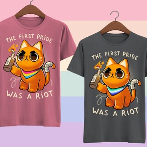 Puede incluir: Dos camisetas con un gato de dibujos animados sosteniendo un c&oacute;ctel Molotov encendido. El texto en las camisetas dice "The First Pride Was A Riot". Una camiseta es rosa y la otra es gris.