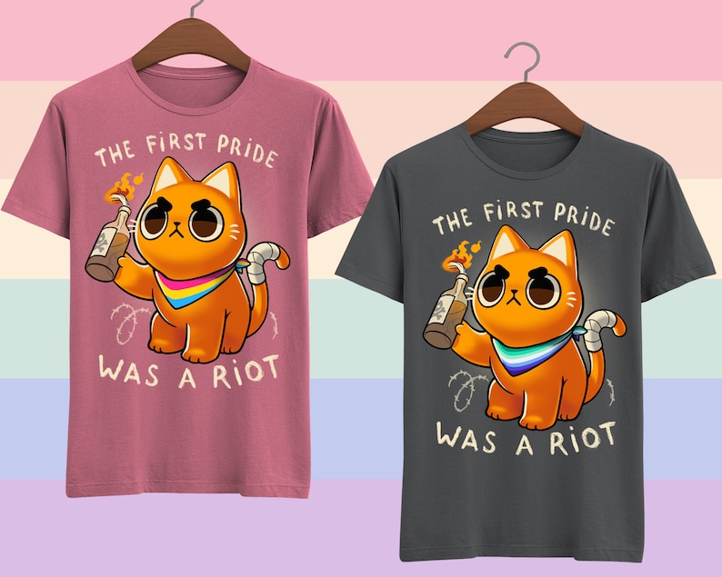 Puede incluir: Dos camisetas con un gato de dibujos animados sosteniendo un c&oacute;ctel Molotov encendido. El texto en las camisetas dice "The First Pride Was A Riot". Una camiseta es rosa y la otra es gris.