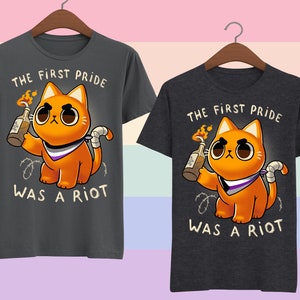 Puede incluir: Dos camisetas grises oscuras con un gato naranja de dibujos animados sosteniendo un c&oacute;ctel Molotov. El gato lleva un pa&ntilde;uelo morado, blanco y negro. El texto en las camisetas dice "The First Pride Was A Riot".