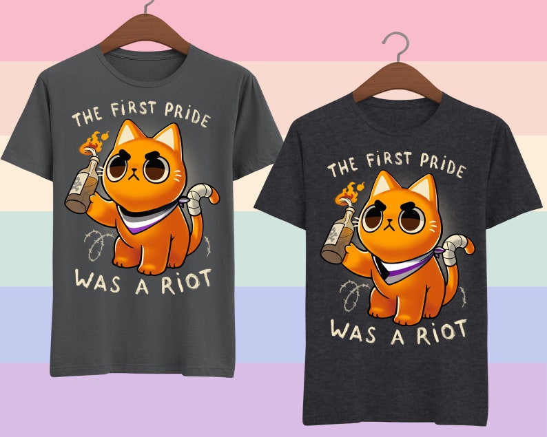 Puede incluir: Dos camisetas grises oscuras con un gato naranja de dibujos animados sosteniendo un c&oacute;ctel Molotov. El gato lleva un pa&ntilde;uelo morado, blanco y negro. El texto en las camisetas dice "The First Pride Was A Riot".