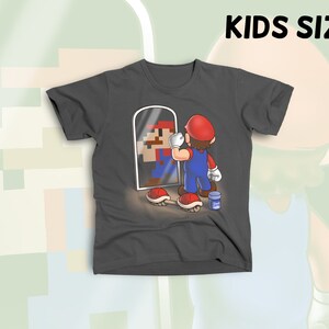 May include: A dark gray t-shirt with a cartoon image of a video game character looking in a mirror. The character is holding a paintbrush and a can of paint. The text "KIDS SIZE" is visible at the top of the image.