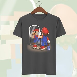 May include: A dark gray t-shirt with a graphic of a video game character looking in a mirror. The character is holding a paintbrush and is surrounded by red and white mushrooms.