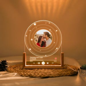 May include: A personalized LED night light with a photo of a couple. The light has a wooden base and a clear acrylic panel with a circular design and star accents. The text "Thinking Out Loud" and a music player interface are visible.