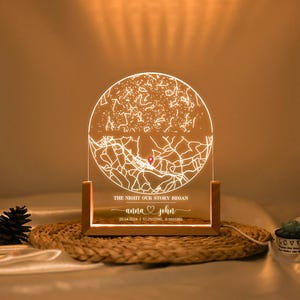 Może przedstawiać: Spersonalizowana lampka nocna z mapą i konstelacjami gwiazd. Światło znajduje się w drewnianej podstawie i zawiera tekst "THE NIGHT OUR STORY BEGAN" oraz imiona "Anna & John" z datą i współrzędnymi.