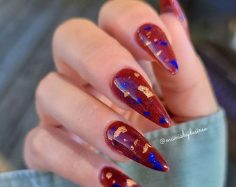 May include: Close-up of a hand with long, pointed nails painted a deep red with gold and blue glitter accents. The nails are styled in a stiletto shape. The text "@manisbydesiree" is visible in the lower right corner of the image.