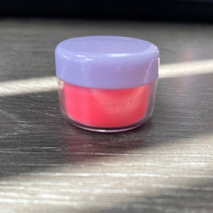 May include: A small, round, clear plastic jar with a purple lid. The jar is filled with a bright pink substance.