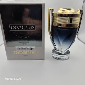 Puede incluir: Un frasco de perfume y su caja. El frasco es de vidrio azul oscuro y transparente con una tapa y detalles dorados. La caja es blanca con la palabra "INVICTUS" y los logotipos de Sephora y Rabanne.