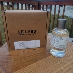 May include: A clear glass perfume bottle with a silver cap and a label that reads "SANTAL 33" sits next to a brown cardboard box. The box has the words "LE LABO" and "GRASSE - NEW YORK" printed on it. The bottle contains 3.4 fl oz of perfume.