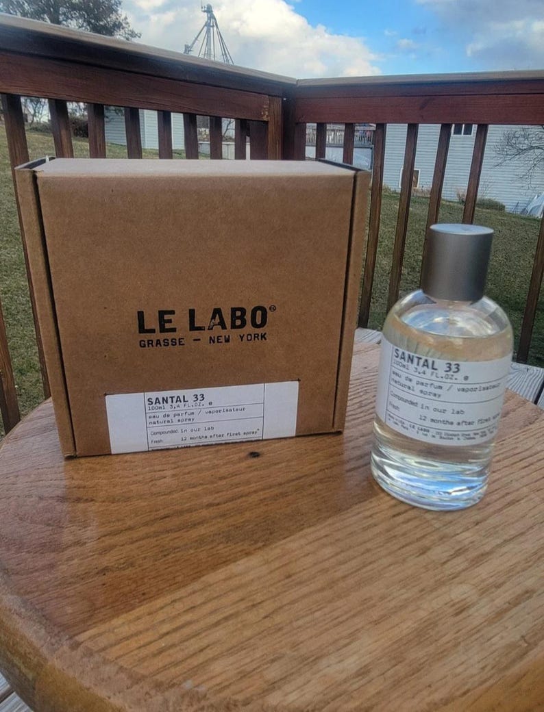 May include: A clear glass perfume bottle with a silver cap and a label that reads "SANTAL 33" sits next to a brown cardboard box. The box has the words "LE LABO" and "GRASSE - NEW YORK" printed on it. The bottle contains 3.4 fl oz of perfume.