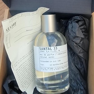 May include: A clear glass bottle of Santal 33 eau de parfum, 3.4 fl oz, with a silver cap, inside a cardboard box. The label includes the text "natural spray." The box contains black tissue paper and a receipt.