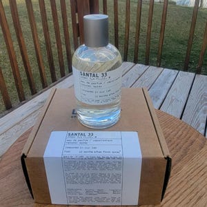 May include: A clear glass bottle of Santal 33 eau de parfum, 3.4 fl oz, with a silver cap, sits atop a cardboard box. The label includes the text "Compounded in our lab" and "12 months after first spray."