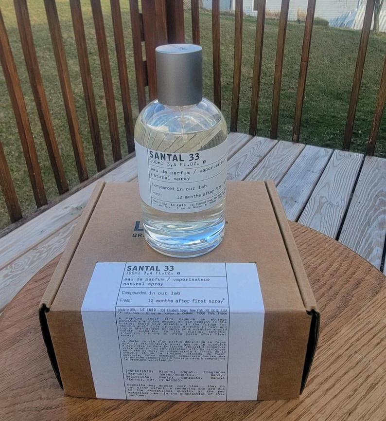 May include: A clear glass bottle of Santal 33 eau de parfum, 3.4 fl oz, with a silver cap, sits atop a cardboard box. The label includes the text "Compounded in our lab" and "12 months after first spray."